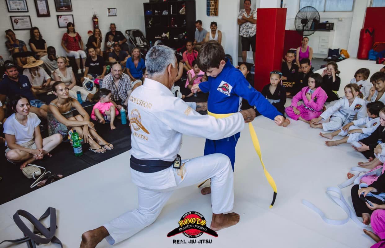 Professor Paulo Guimaraes tying a new belt on a young student while families watch